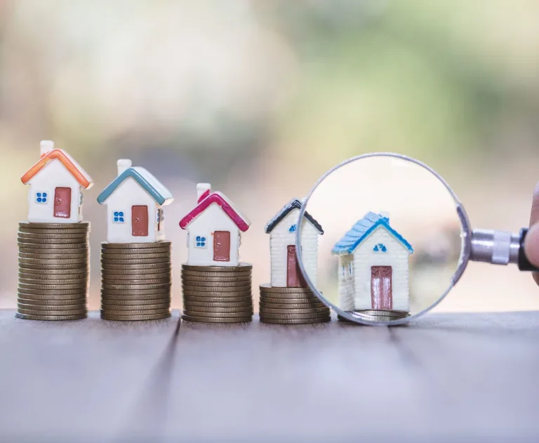 This image shows several small house figurines stacked on piles of coins, with each stack getting progressively shorter. The last, and shortest, stack has a magnifying glass next to the house.