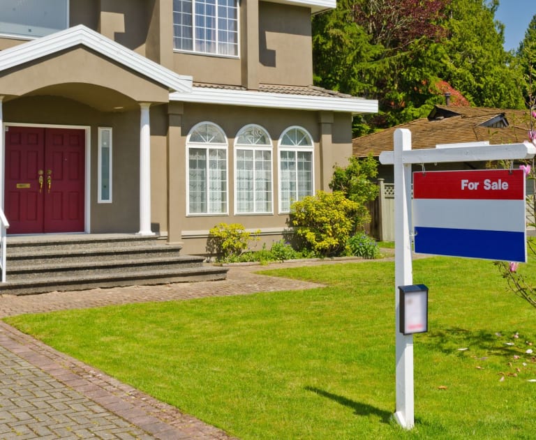 A red and blue for sale sign, posted in front of a tan house with white trim and red door.