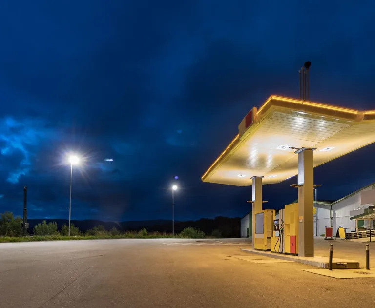 The image features a lighted canopy over fuel pumps with a dark, blue sky in the background.