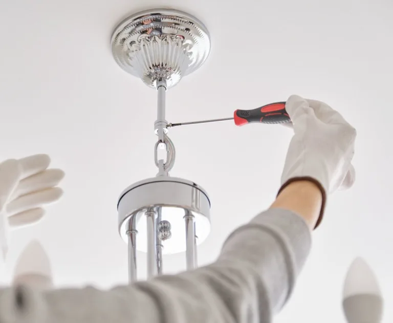 This image shows the installation or repair of a chandelier light fixture. A person is using a screwdriver to secure a polished metal chandelier to the ceiling canopy.