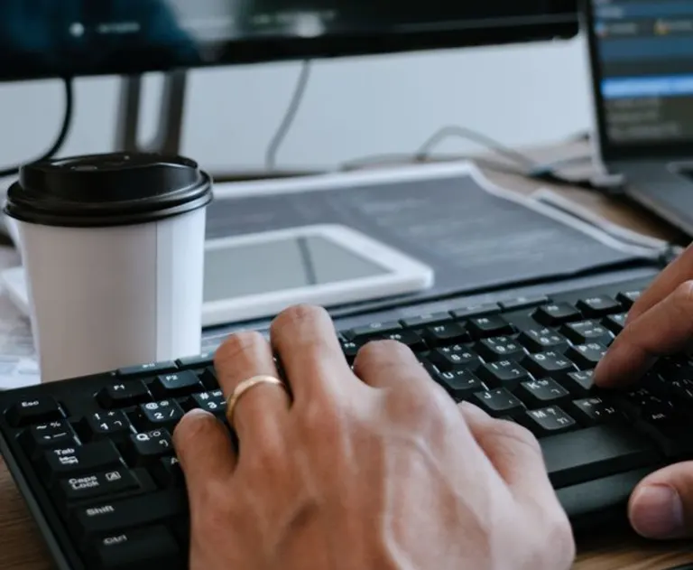 This image shows a person typing on a black computer keyboard, likely in an office setting. The keyboard appears to be a standard QWERTY layout with numeric keypad. In the background, there is a cup of coffee and additional monitors. The overall scene is a typical workspace for tasks like coding, data entry, or general office work.
