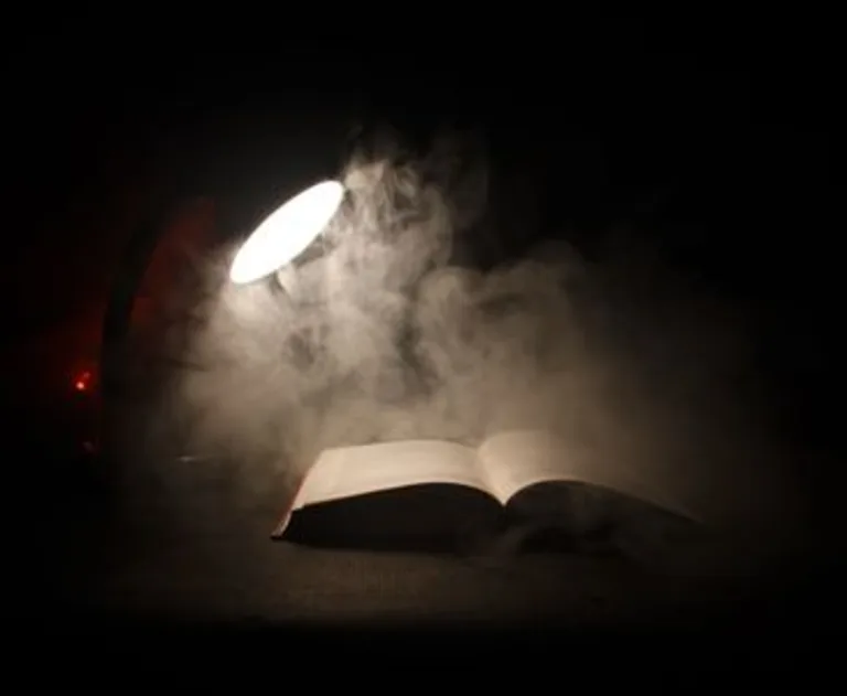 This image depicts a surreal scene featuring an open book illuminated by a glowing desk lamp on a dark background, surrounded by wisps of smoke.