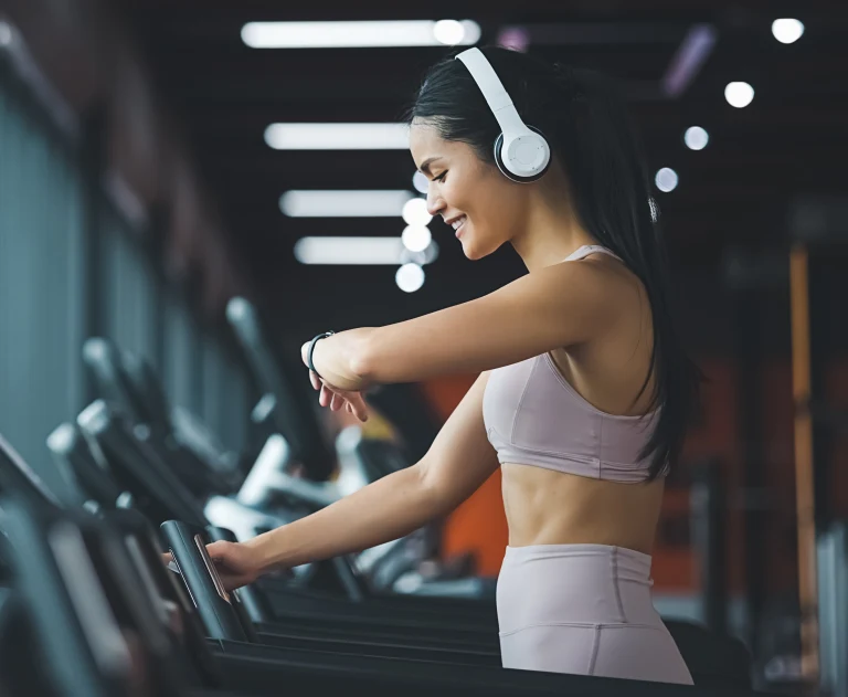 A woman stands at the treadmill in a gym. She is wearing cream colored headphones, a light pink sports top and matching shorts, and smiling at her smart watch.
