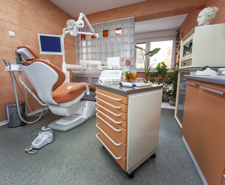 This image shows a modern dental office treatment room designed for patient comfort. It features a reclining dental chair and a mobile instrument trolley. The room includes specialized lighting equipment and storage cabinets. The overall design aims to create a soothing environment with soft colors and natural light.