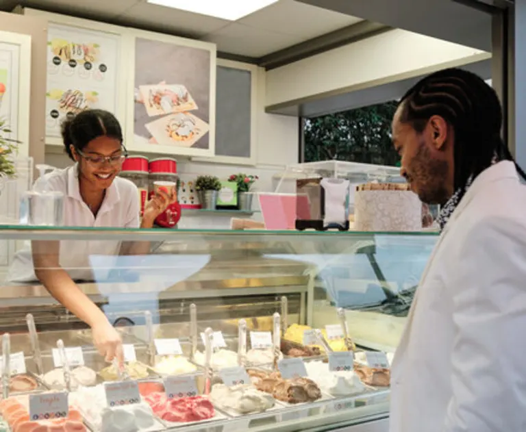 This image shows a customer selecting a flavor at an ice cream shop. A smiling employee is assisting the customer behind a glass display counter. The counter features a variety of gelato or ice cream flavors in containers. The scene depicts a typical interaction in a commercial dessert parlor.