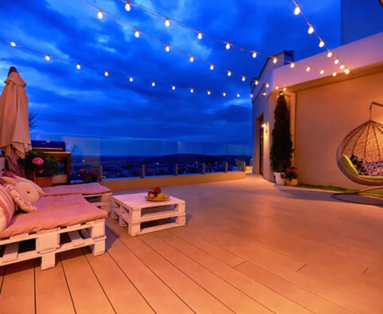 This image shows a cozy rooftop patio area featuring a lounge zone, a hanging egg chair, and string lights during a warm summer evening.