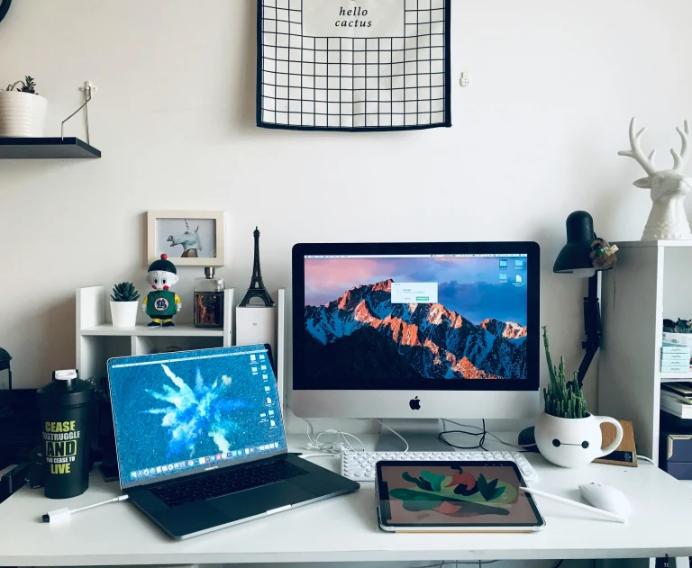 This image displays a modern home office setup featuring a clean white desk, a laptop, an iMac, and a tablet.