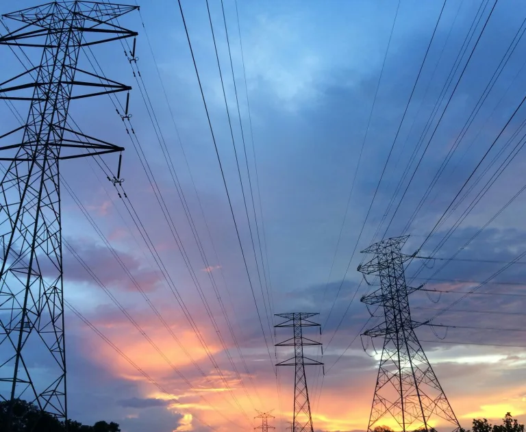 The image displays high-voltage transmission towers and power lines against a sunset sky. These towers support electrical cables designed to efficiently transmit large amounts of power over long distances.