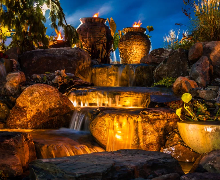 This image displays a waterfall fountain landscape designed for evening ambiance by using landscape lighting.