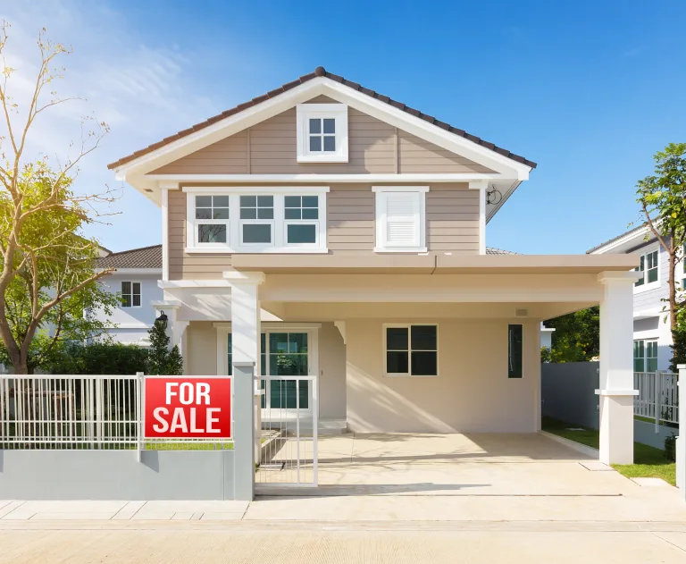 This image shows a modern two-story residential house for sale, featuring a light brown and white exterior, a covered carport, and a white metal fence with a "For Sale" sign attached.