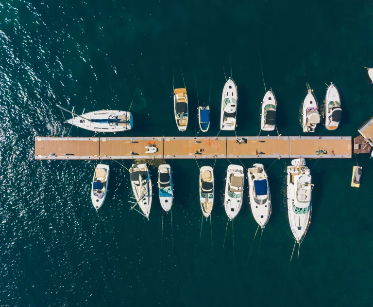 This image shows an aerial view of various yachts and sailboats moored at a dock in a marina.