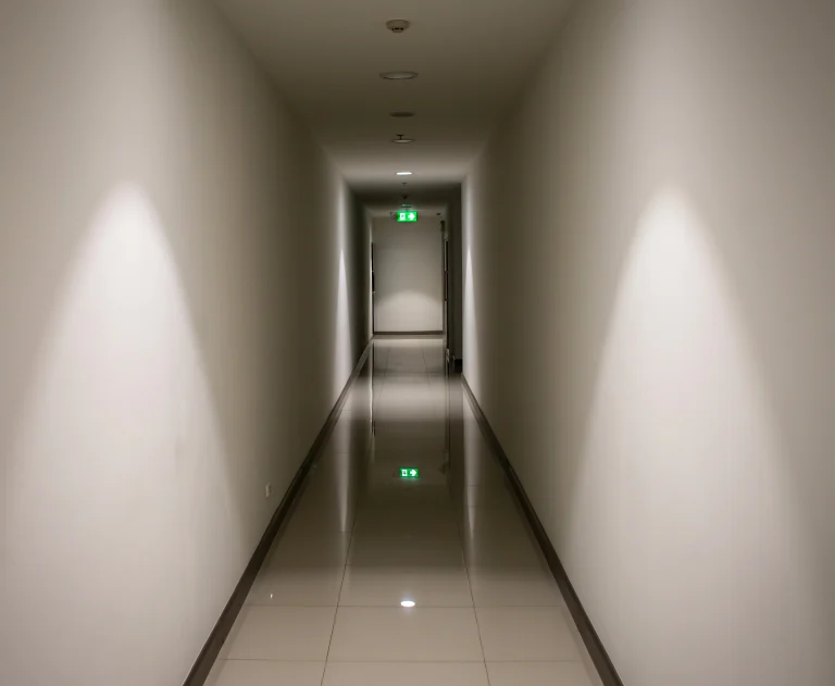 This image shows a long, empty corridor, likely inside a modern hotel or commercial building, featuring polished floors and illuminated exit signs.