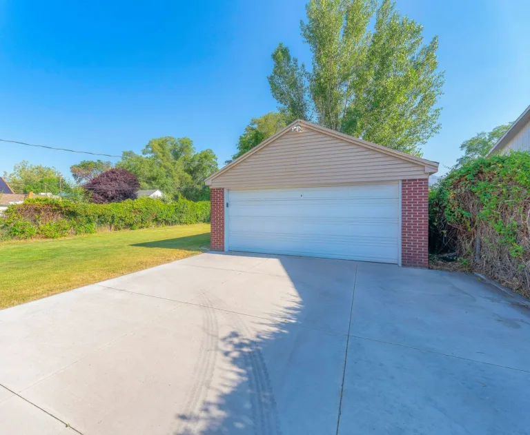 This image features a two-car garage with a concrete driveway approach. The structure has a brick and beige siding exterior with a white garage door. A mature tree stands behind the garage, and there is a fence covered in vines on the right. The property includes a grassy lawn area next to the driveway.