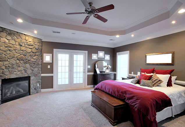 This image features a cozy bedroom with a stone-accented fireplace on one side and recessed lighting.