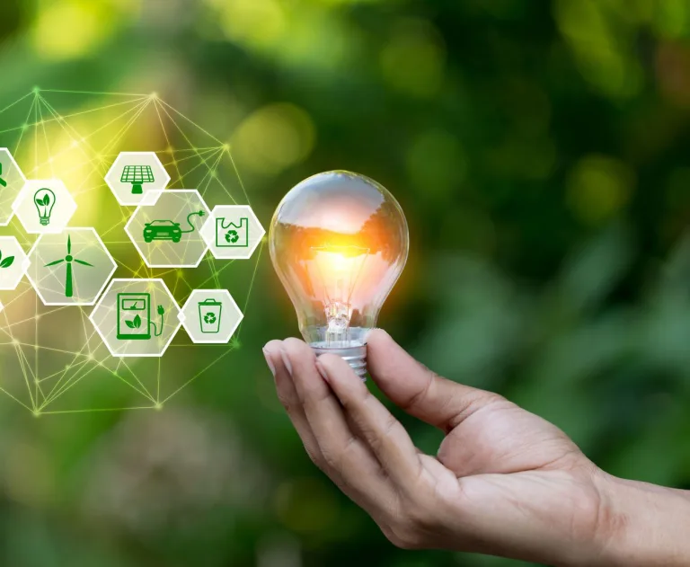 This image represents a concept focused on renewable energy and environmental sustainability, featuring a hand holding a glowing light bulb surrounded by interconnected green energy icons.