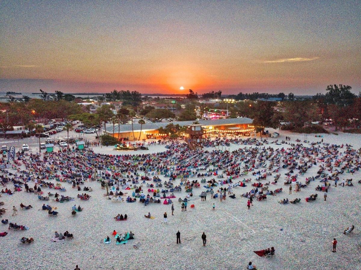 This image shows a large crowd gathered around a stage on a Florida beach during sunset.