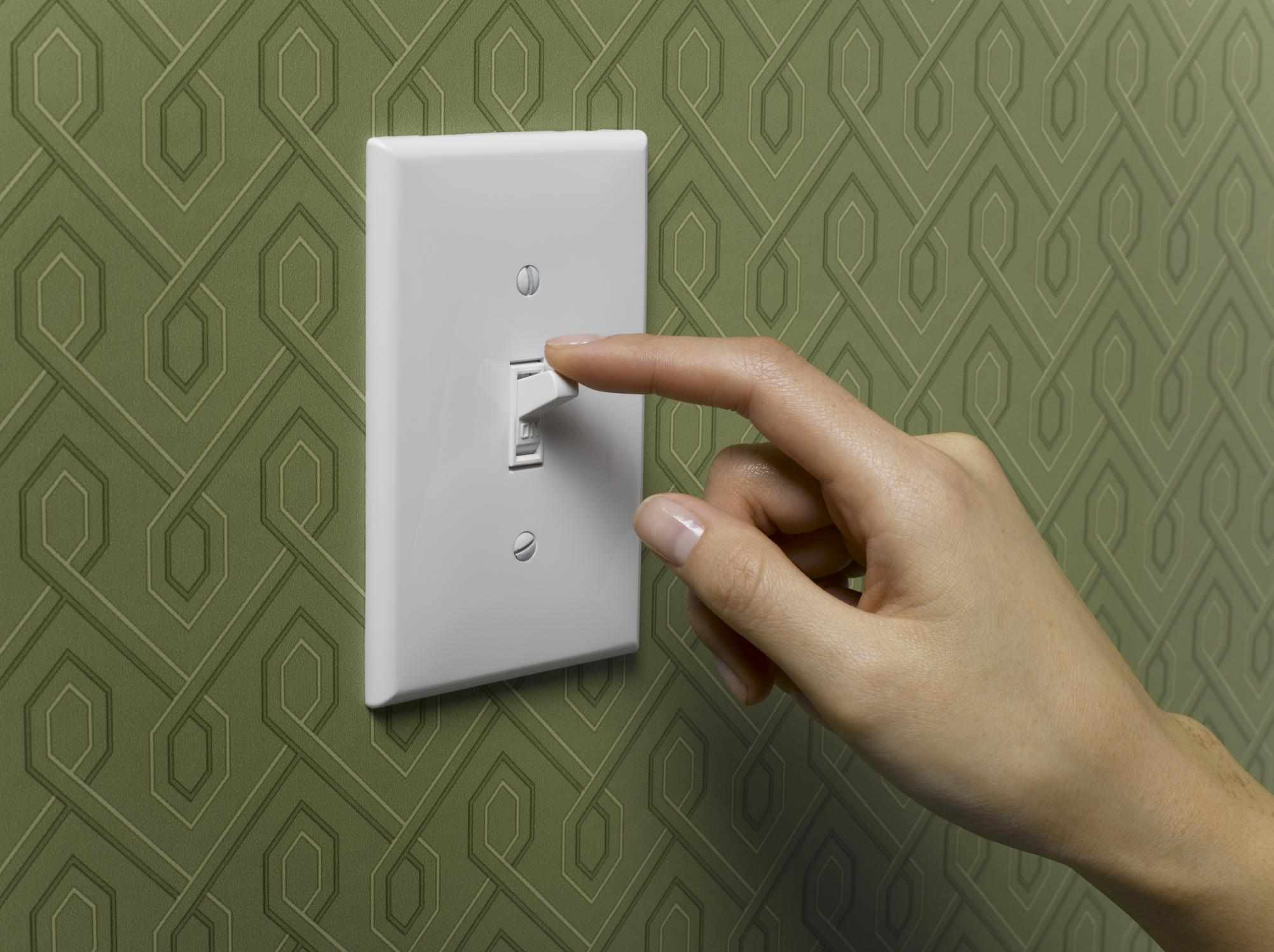 The image shows a standard white toggled light switch mounted on a wall with green patterned wallpaper.