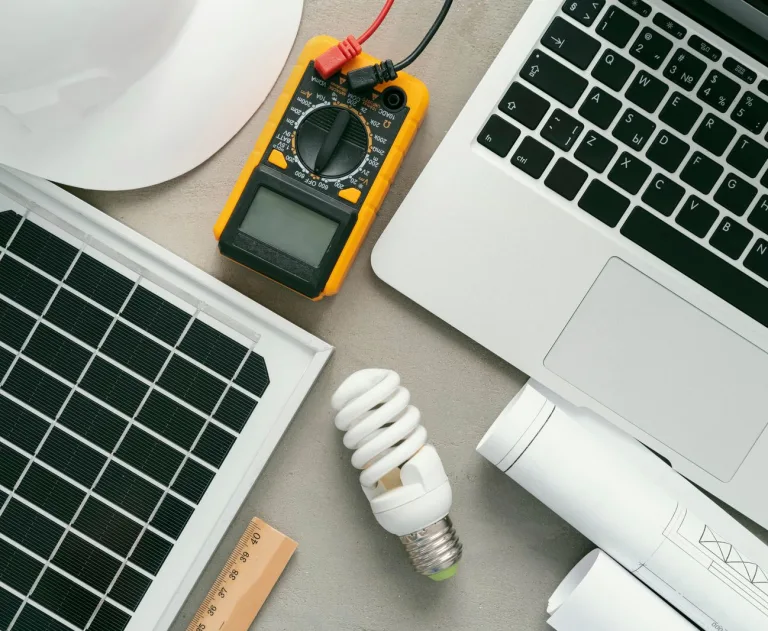 This image displays essential equipment for a solar power project, including a photovoltaic panel, a digital multimeter for testing circuits, a laptop for monitoring or design, and a light bulb.