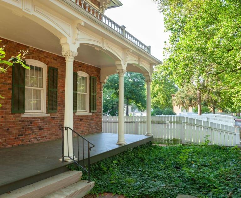 This image shows a historic brick house with a covered front porch, a white picket fence, and lush green landscaping.