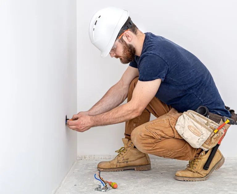 The image depicts a professional electrician performing electrical installation work on a white wall.