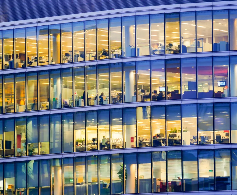 This image depicts the exterior of a modern office building with large, glass-paned windows. The interior offices are illuminated, showing individual workstations, desks, and office equipment.