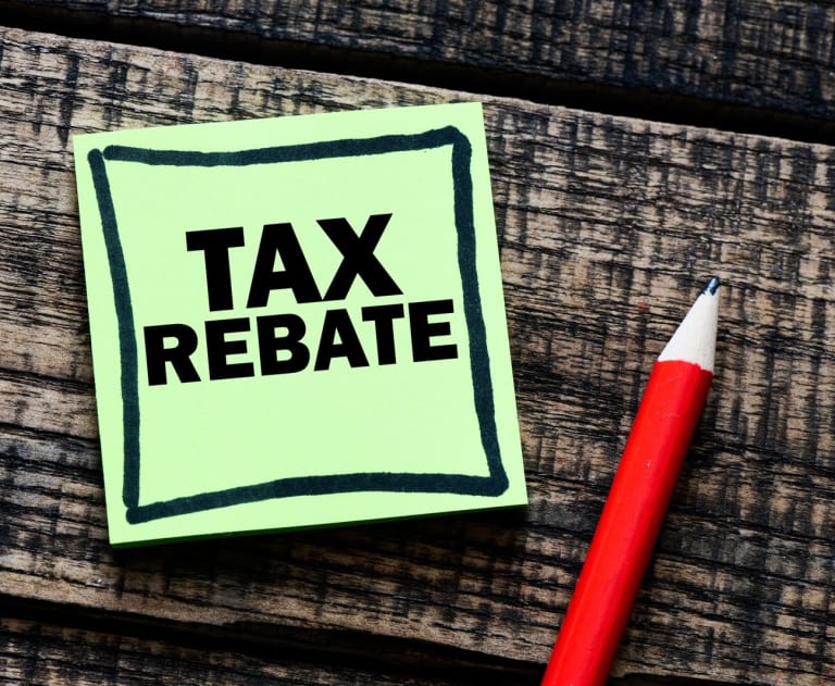 The image features a light green square sticky note positioned on a dark, rustic wooden surface.The sticky note is outlined with a bold, hand-drawn black border. Written inside the border are the words "TAX REBATE" in large, uppercase, sans-serif black typography. Resting diagonally to the right of the note is a sharpened red wooden pencil with a exposed light wood tip and a sharp black graphite point. The underlying surface consists of textured, dark brown wooden planks with distinct grain lines and weathering.