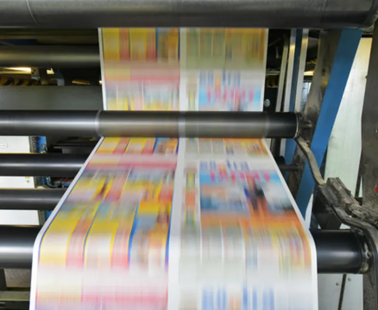 This image depicts a high-speed printing press in operation, likely a web offset machine based on the continuous roll of paper shown.