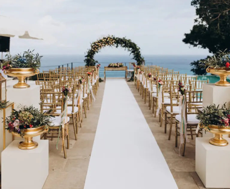 An outdoor wedding setup at features gold chairs and a white aisle leading to a floral arch against a scenic ocean backdrop, flanked by lush greenery in gold urns.