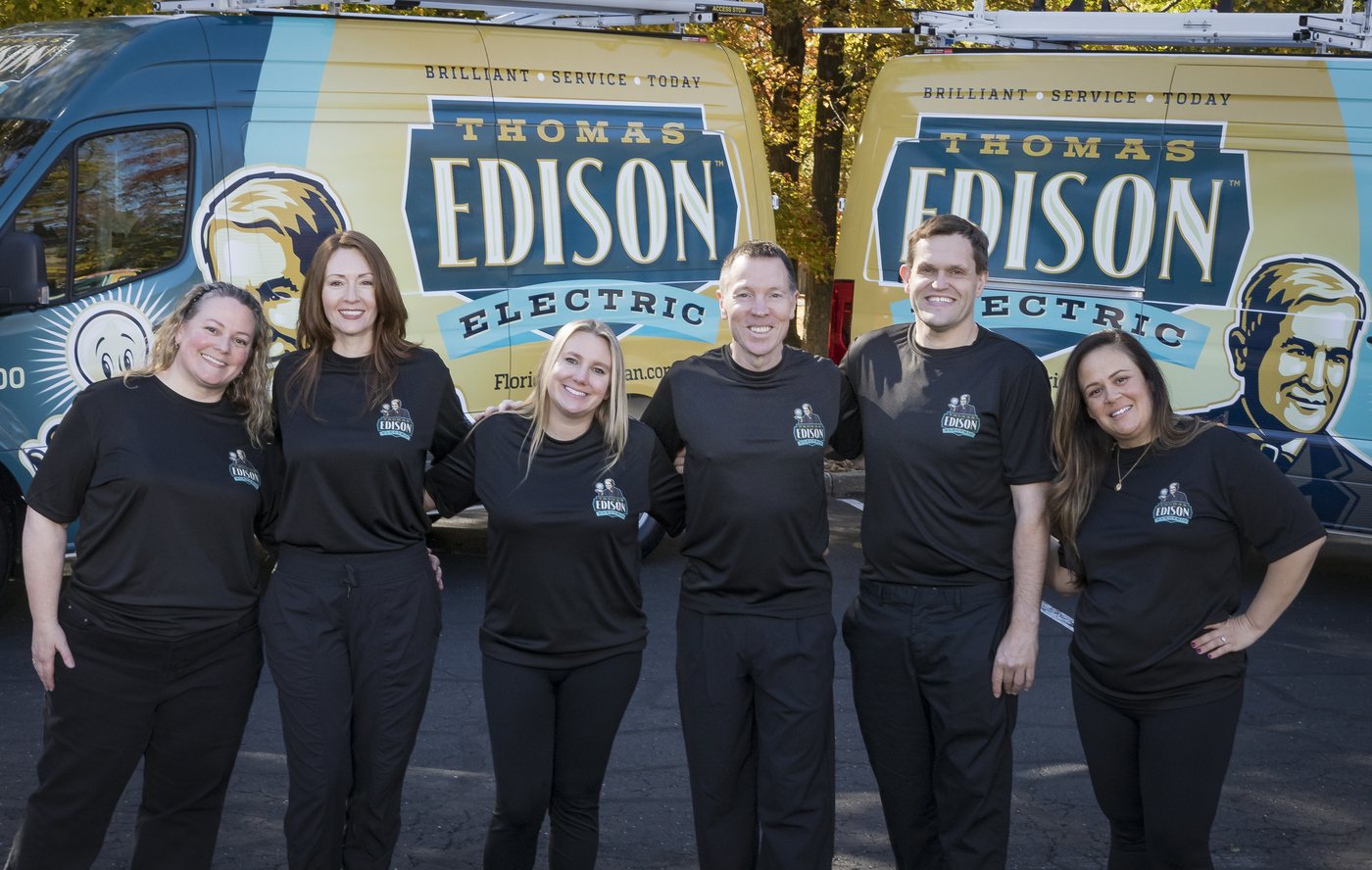 tee-fl-team-hero - Thomas Edison Electric Six Edison Electric team members in black shirts stand arm in arm in front of branded service vans outdoors, smiling at the camera, with 'Thomas Edison Electric' logos visible on the vehicles.